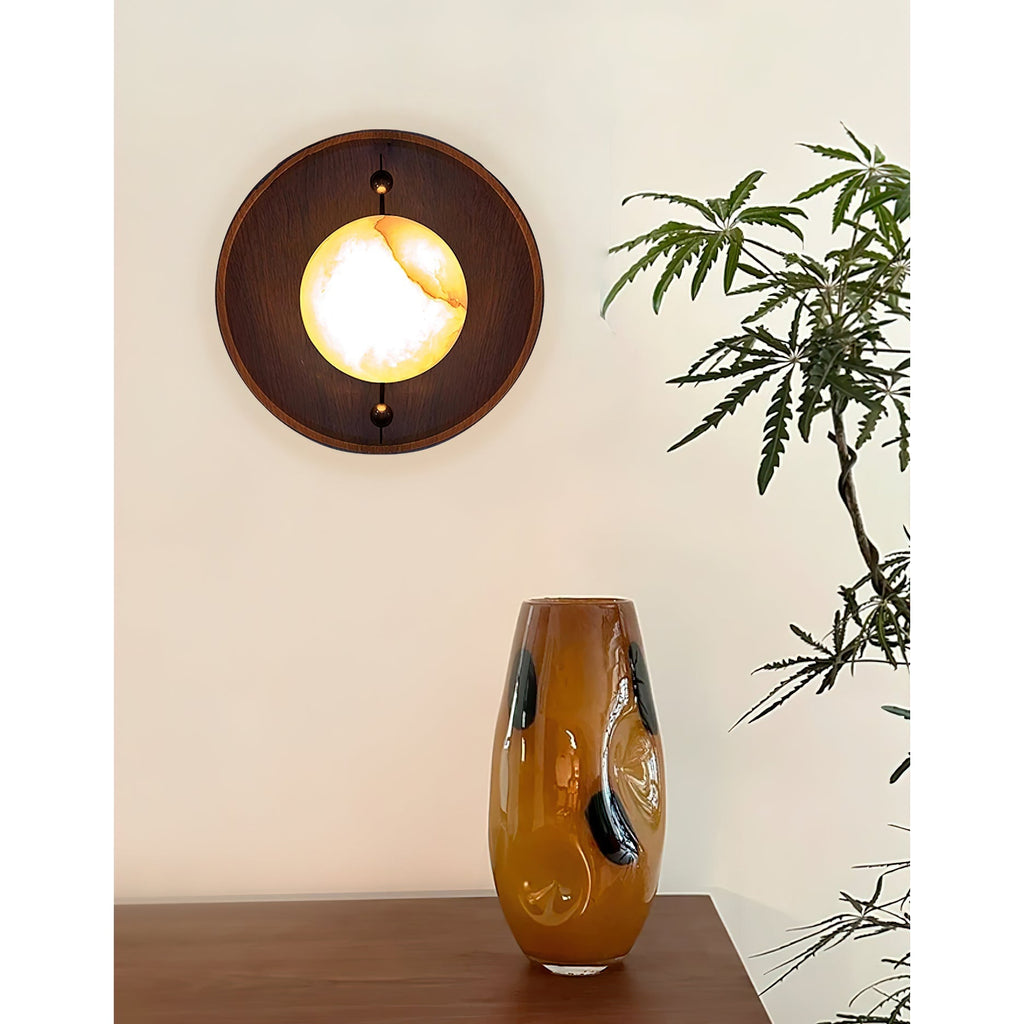 The round Clérachic Alabaster Wall Sconce lit beside an amber glass vase, its stone glowing white at the centre and gold at the edge.