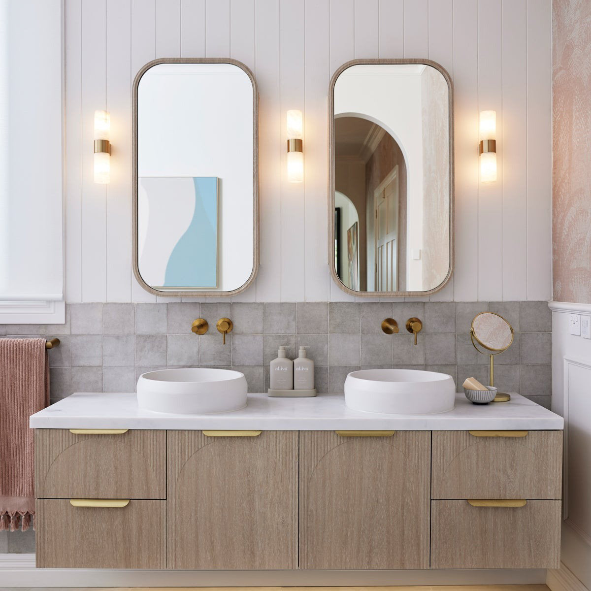 Clérachic Alight sconces lit between two arched mirrors over a double oak vanity with white stone basins.