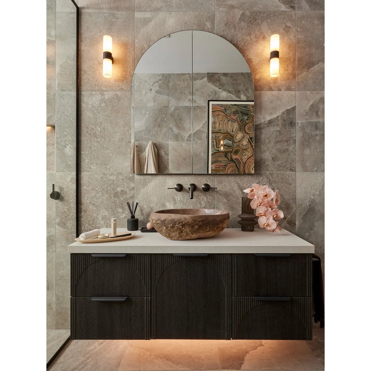 The Clérachic Alight sconces in black, lit either side of an arched mirror in a stone-tiled bath with a carved basin.
