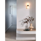 Clérachic Alight sconce lit above a curved plaster ledge in a hallway, its glow washing the wall beside a dried stem.
