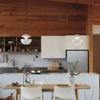 Two white Clérachic pendants hang over a wooden table in a kitchen with white tiles, open shelves and a timber wall.
