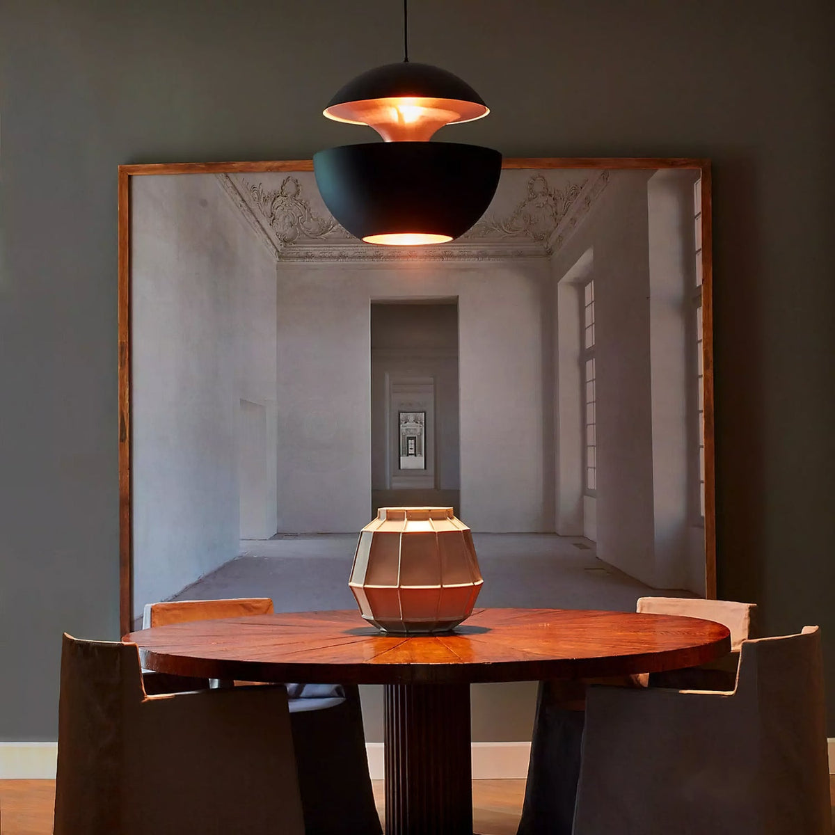 Black pendant with a gold interior above a round wooden table, set against a dark wall and a large framed picture, from Clérachic.