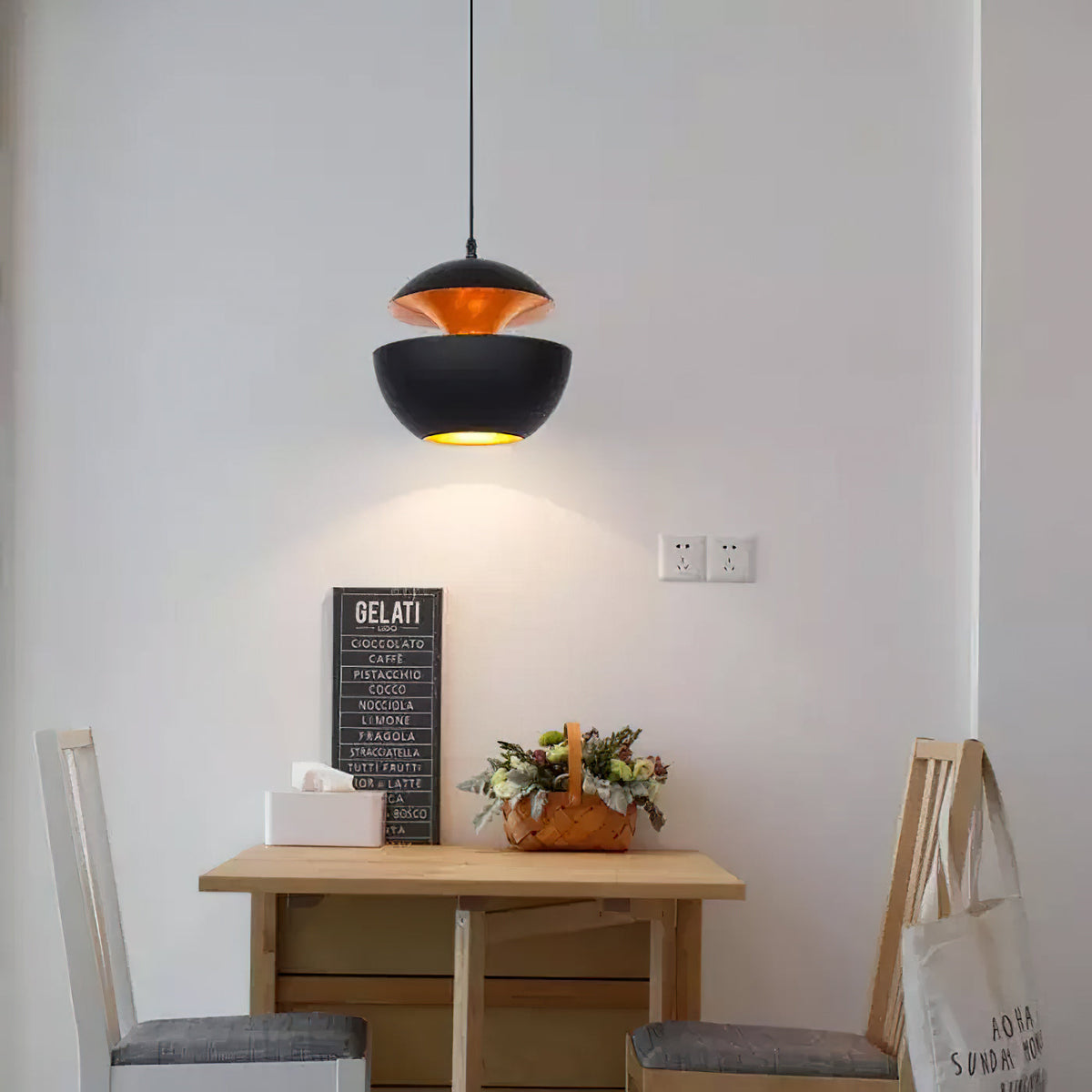 Black bowl pendant with a gold interior above a small wooden table and a sign, against a plain white wall, from Clérachic.