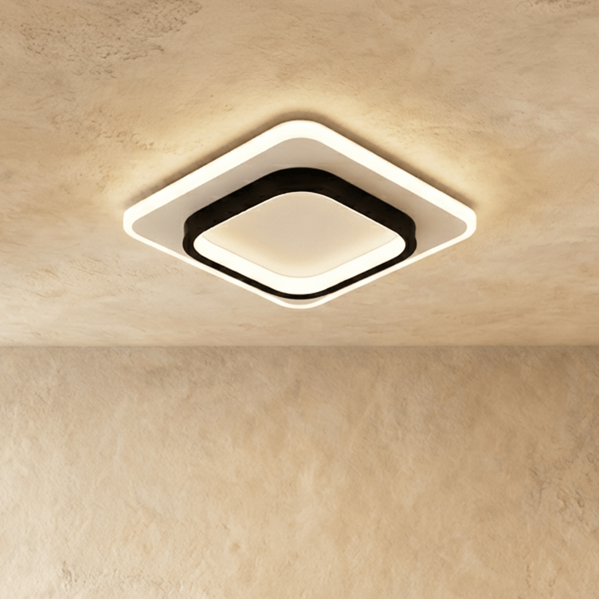 Clérachic rounded-square ceiling light with a black inner frame and white acrylic panel, glowing warm on a plastered ceiling.
