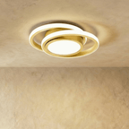 Clérachic ceiling light with two offset white rings and a gold-finish core, glowing softly on a plastered ceiling.