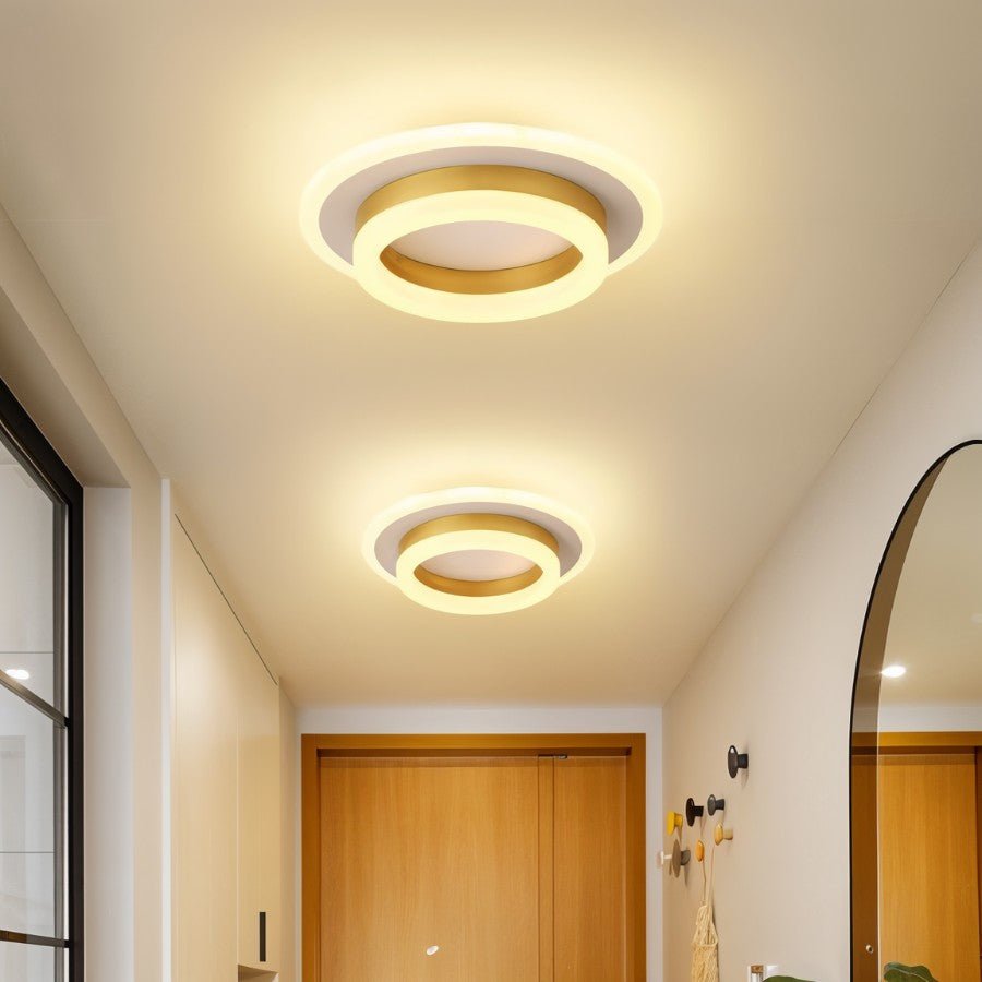 Two Clérachic round gold-finish ceiling lights in graduated sizes, lighting a cream hallway with a timber door and arched mirror.