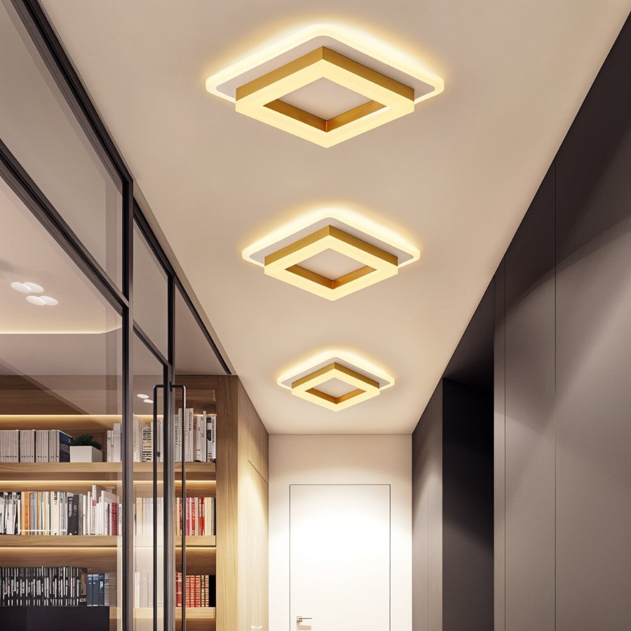 Three Clérachic square gold-finish ceiling lights in graduated sizes, lighting a long corridor beside a glazed bookcase wall.