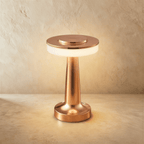 Clérachic cordless table lamp in polished rose gold, lit, its flat disc head above a softly glowing opal band
