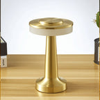 Clérachic cordless table lamp in brushed gold on a wooden desk with books and plants, disc head over an opal band