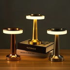 Three Clérachic cordless table lamps in rose gold, gold and silver glowing on a wooden shelf around a stack of books