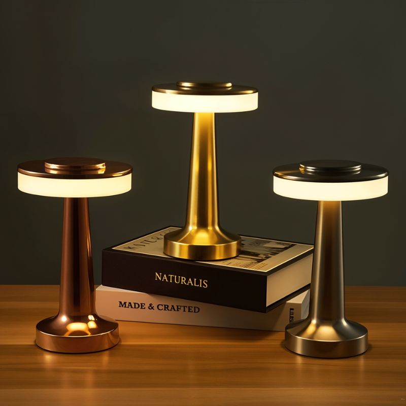 Three Clérachic cordless table lamps in rose gold, gold and silver glowing on a wooden shelf around a stack of books
