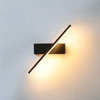 Clérachic black bar wall light rotated to a diagonal, its warm lit arm crossing a black rectangular backplate on a pale wall.