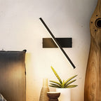 Clérachic black bar wall light angled across its plate on a speckled plaster wall, beside a bedside cushion and potted plant.