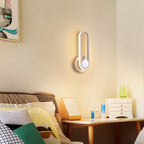 Clérachic white oval ring wall light at a bedside, glowing softly beside a bed with layered cushions and a small chest.