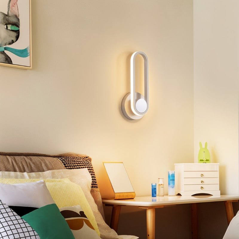 Clérachic white oval ring wall light at a bedside, glowing softly beside a bed with layered cushions and a small chest.
