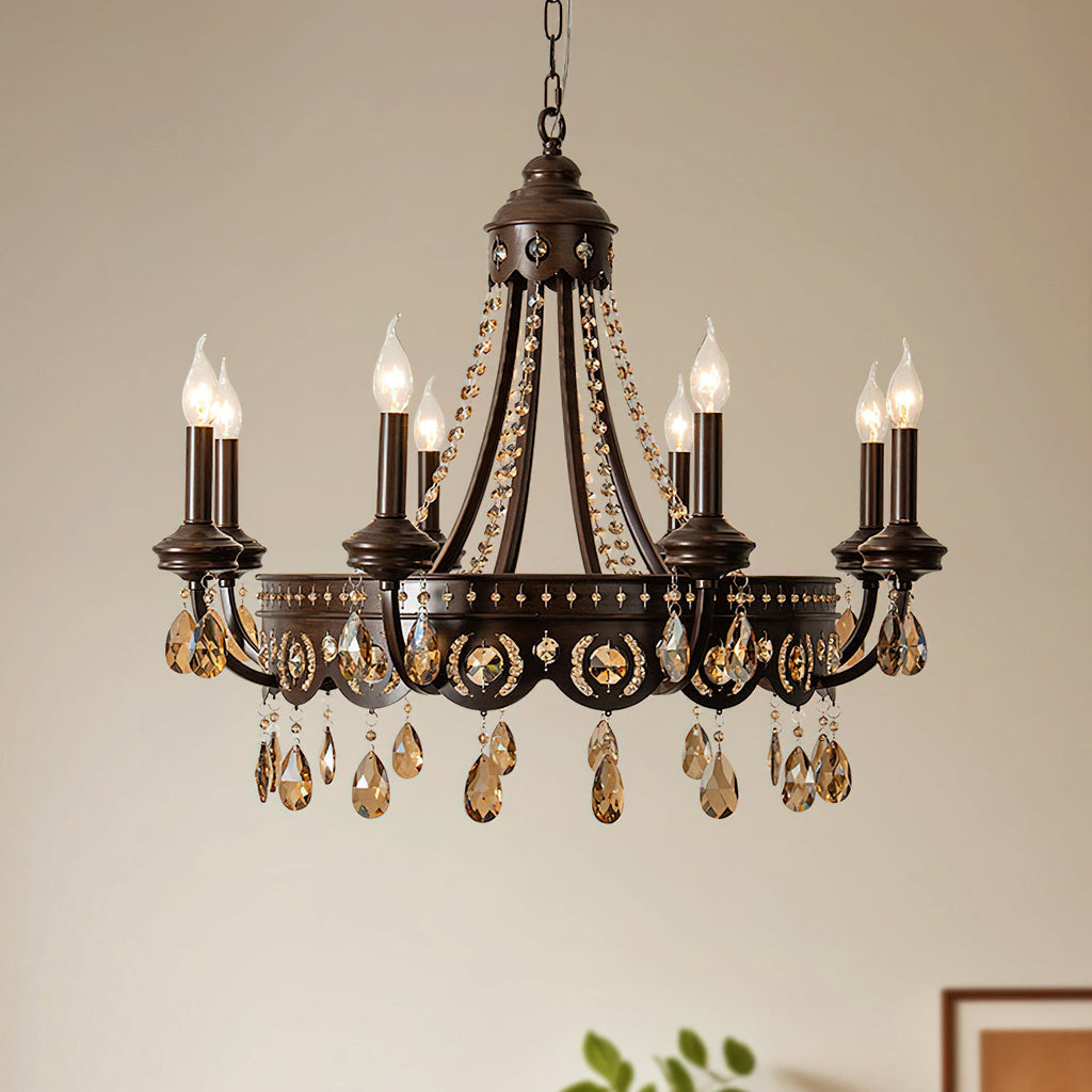 A lit Clérachic chandelier with candle-style bulbs and amber crystal drops, hung against a warm cream wall with a plant below.