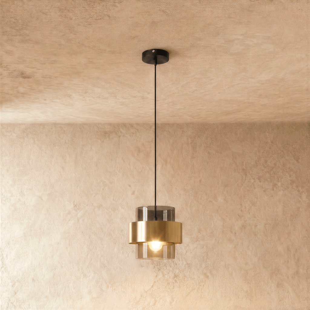 The Clérachic pendant in smoky gray glass with a brass band, hanging from a black cord against a soft plaster wall.