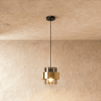 The Clérachic pendant in smoky gray glass with a brass band, hanging from a black cord against a soft plaster wall.