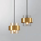 Two Clérachic cylinder pendants side by side, brass-banded glass shades glowing warm white against a plain grey background.