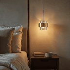 The Clérachic cylinder pendant glowing warm white beside a wood nightstand with a mug and a book on top.
