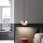 The Clérachic smoky glass pendant with a brass band glowing warm white above a low stool beside a tall window.