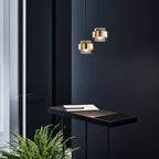 Two Clérachic cylinder pendants with brass bands glowing warm white above a dark desk with a notebook and pen.