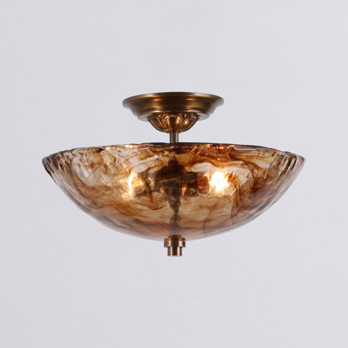 Lit, this Clérachic ceiling light shows bulbs glowing through its marbled amber glass bowl, with a bronze canopy and finial on a pale grey backdrop.