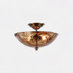 This Clérachic ceiling light seen from slightly below on a pale backdrop, its bronze stem and canopy rising above the lit amber glass bowl.