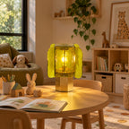 Green and gold Clérachic table lamp glowing on a round wooden children's table, with a picture book and a toy rabbit beside it.