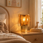 Amber glass Clérachic table lamp on a wooden bedside chest, with a toy giraffe, stacked books and a cream curtain behind.