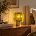 Green-glass Clérachic table lamp lit on a walnut chest, with a stone bowl and books, beside a bed with a cushion.