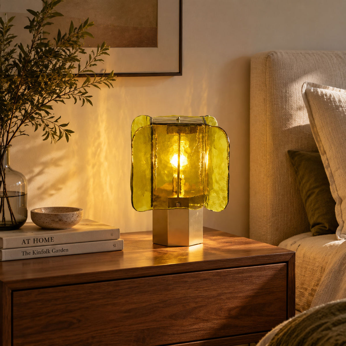 Green-glass Clérachic table lamp lit on a walnut chest, with a stone bowl and books, beside a bed with a cushion.