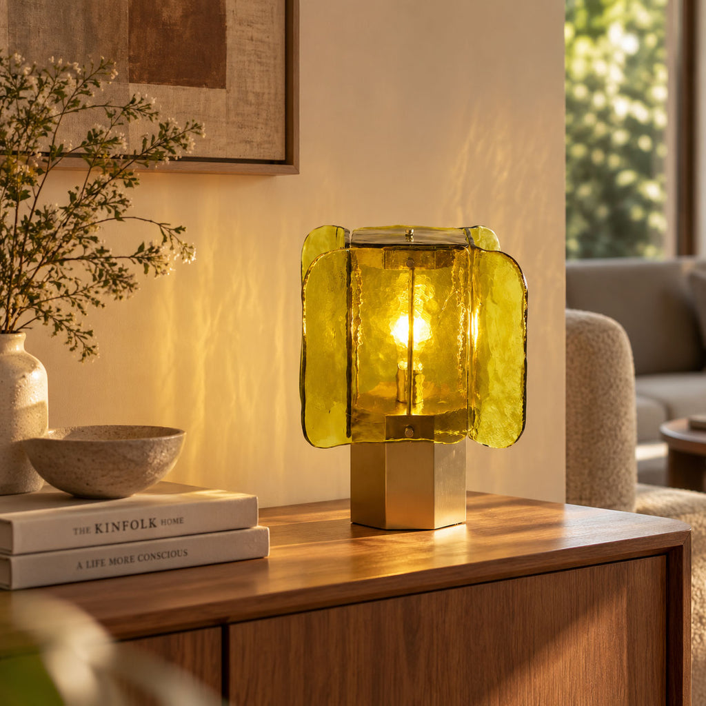 Lit green Clérachic table lamp on a wooden sideboard, with a stone bowl, books and a vase of leafy stems in warm light.