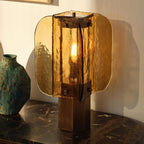 Close view of the amber glass panels and bronze-toned base of this Clérachic table lamp, on a dark marble ledge beside a teal vase.