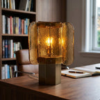 Amber glass Clérachic table lamp lit on a walnut desk, in front of a bookcase, with an open notebook and a leather chair.