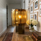 Large amber Clérachic table lamp on a rustic console in a hallway, beside white hydrangeas and a wall of framed photographs.