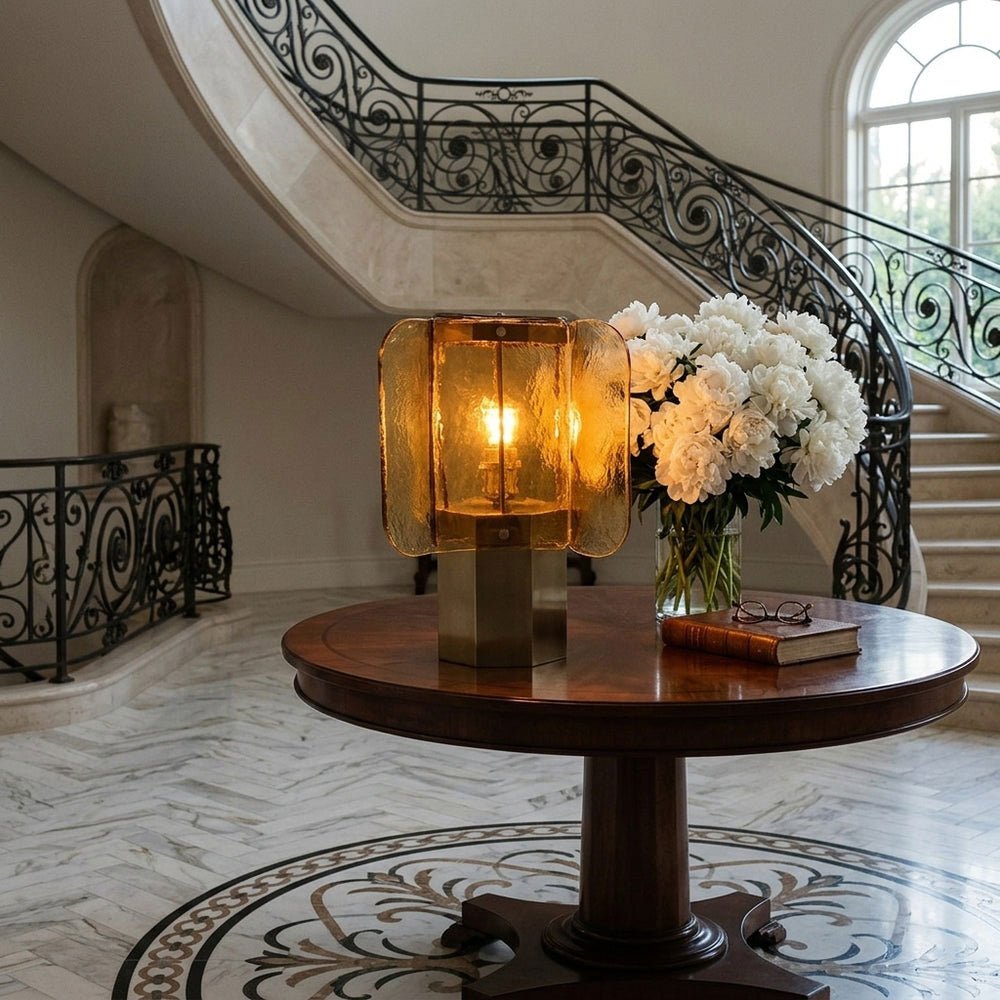 Amber glass Clérachic table lamp on a round wooden table in a hallway with a sweeping stair and a vase of white flowers.