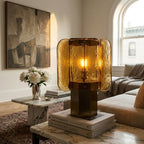 Lit amber Clérachic table lamp on a stack of books on a marble table, with an arched window and a sofa behind.