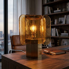 Amber glass Clérachic table lamp lit on a walnut desk beside a leather notebook, with a bookcase and city view behind.