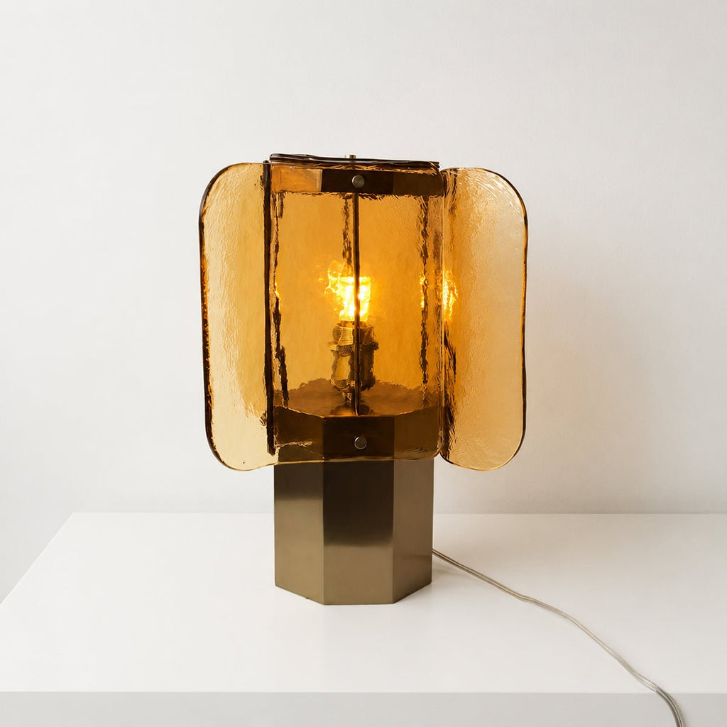 Front view of a lit amber Clérachic table lamp on a white surface, its warm bulb glowing behind four wavy glass panels.