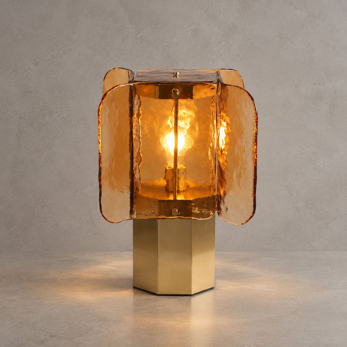 Lit amber glass Clérachic table lamp on a brass base, casting a warm pool of light on a stone-grey surface.