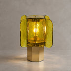 Green and gold Clérachic table lamp lit on a brass base, its chartreuse glass panels glowing against a stone-grey backdrop.