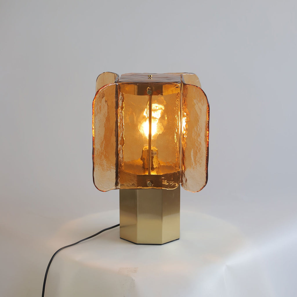 Amber glass Clérachic table lamp lit on a draped white surface, with a black cord trailing from the brass octagonal base.