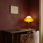 Clérachic table lamp glowing on a walnut console desk against a deep burgundy wall, beside a laptop and a floral armchair.