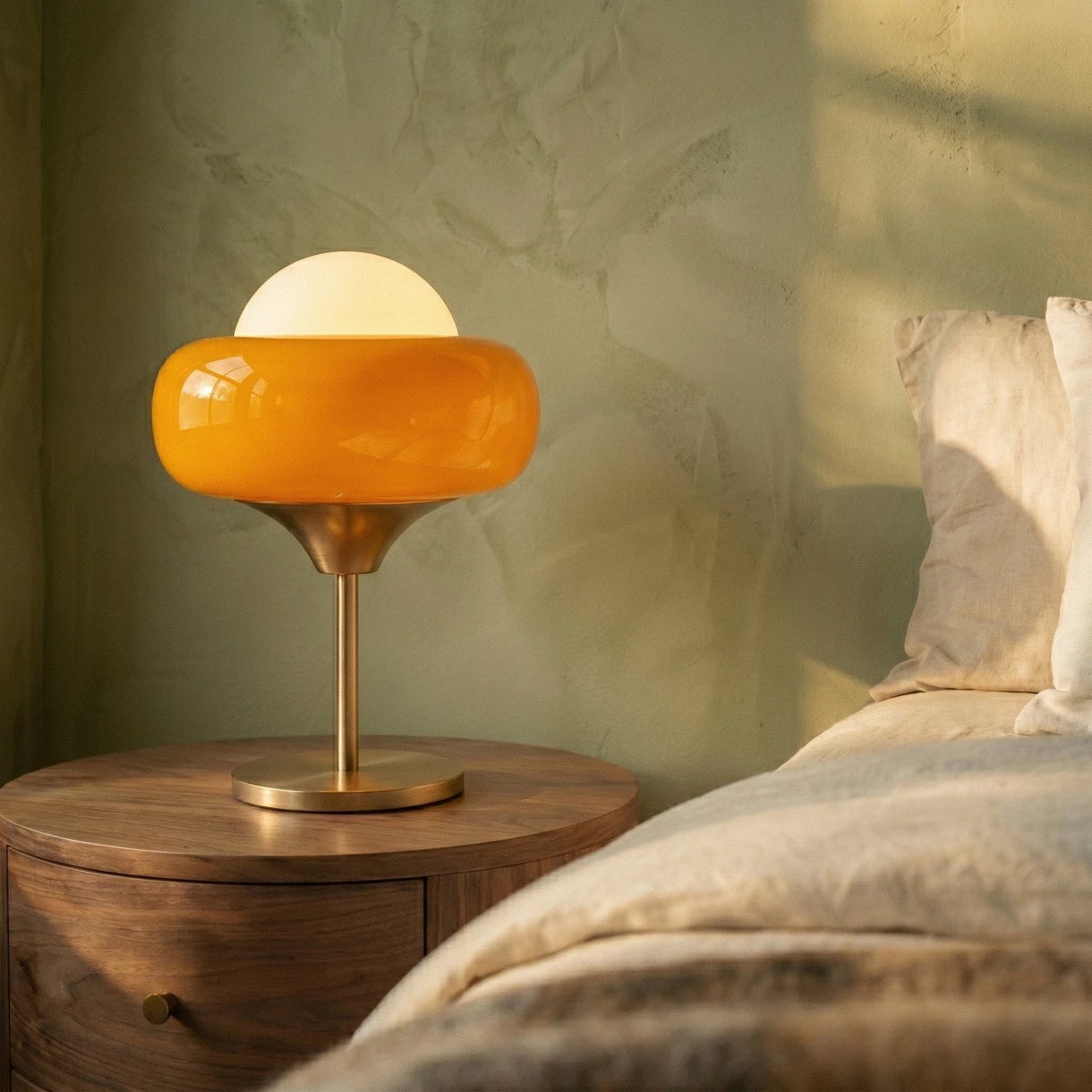 Clérachic table lamp with an amber glass shade and a brass-toned stem and base on a walnut bedside unit by a green wall.