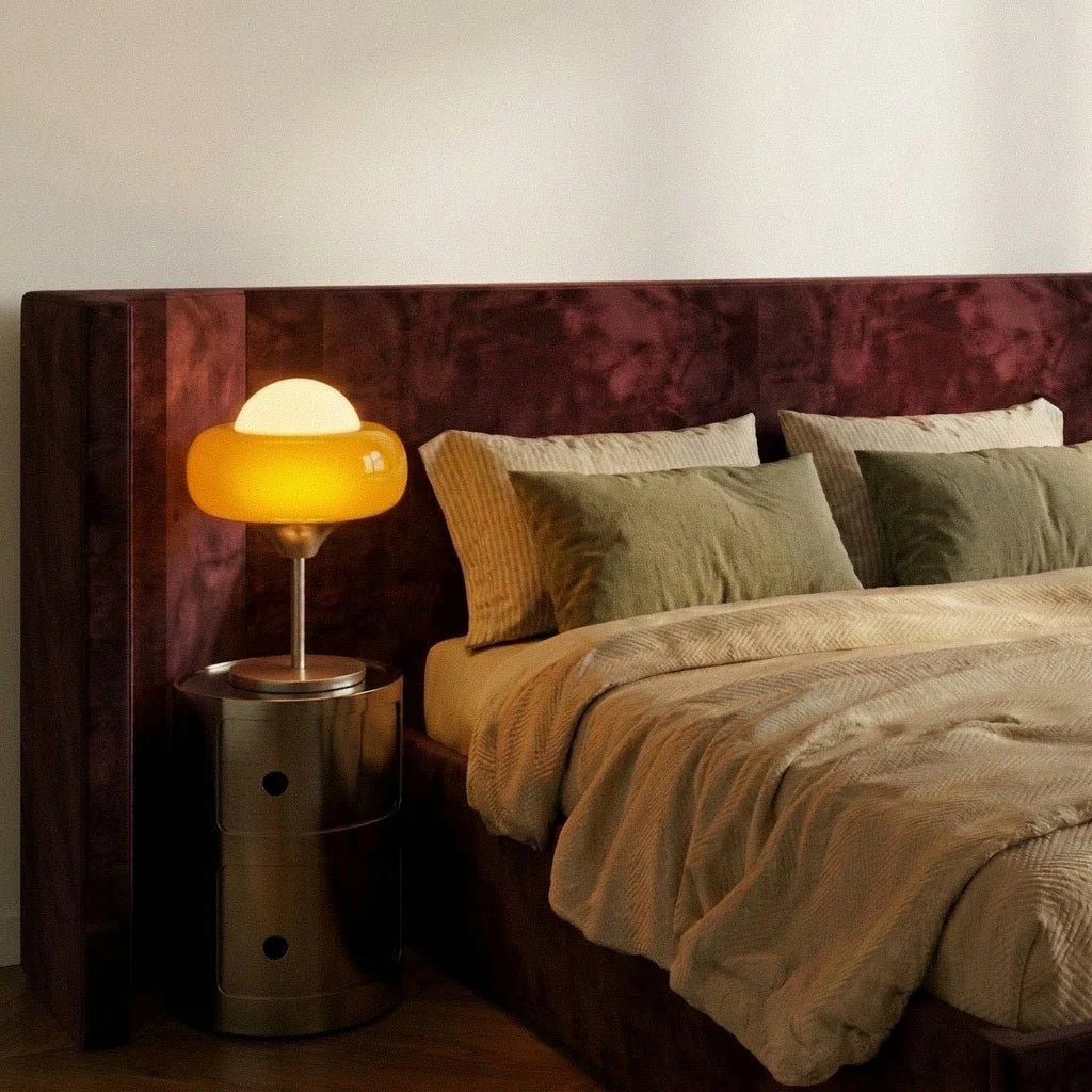 Clérachic table lamp lit beside a burgundy velvet headboard, standing on a chrome round bedside unit with striped linen bedding.