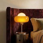 Clérachic amber glass table lamp glowing on a polished metal round bedside unit against a plum velvet headboard and cream wall.