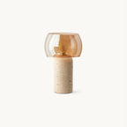 A Clérachic table lamp with an amber glass shade on a travertine column base, shown on a plain white backdrop.