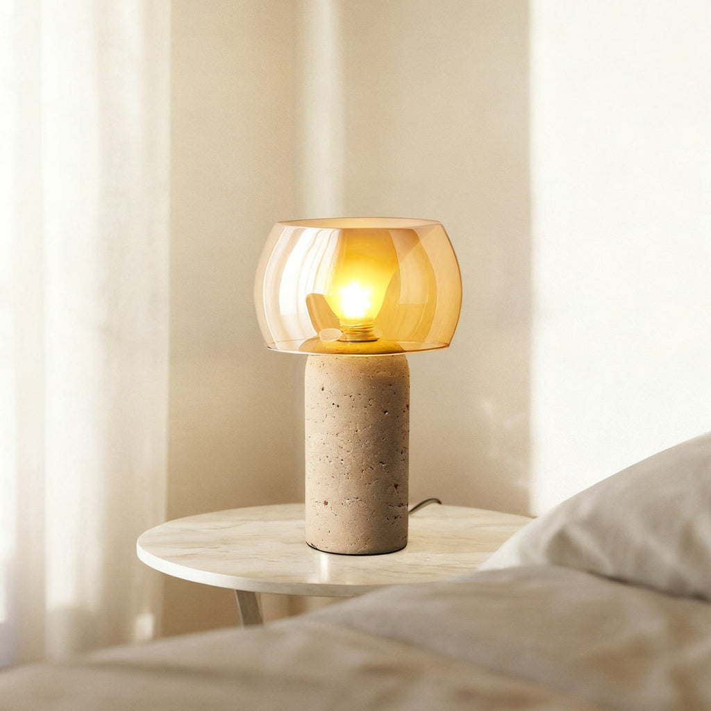 Lit Clérachic table lamp with an amber glass shade glowing beside a bed, on a small round marble-topped side table by a sheer curtain.