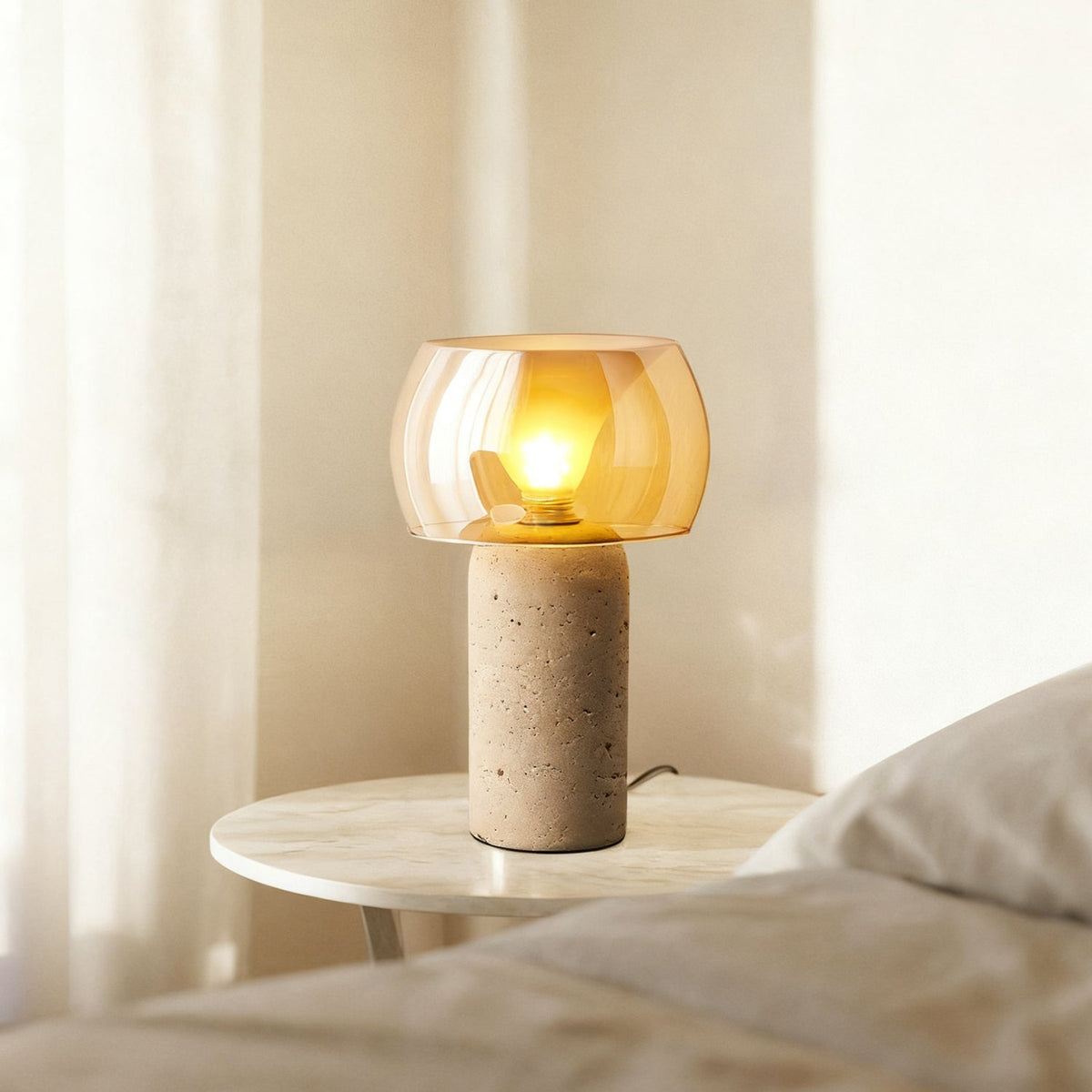 Lit Clérachic table lamp with an amber glass shade glowing beside a bed, on a small round marble-topped side table by a sheer curtain.